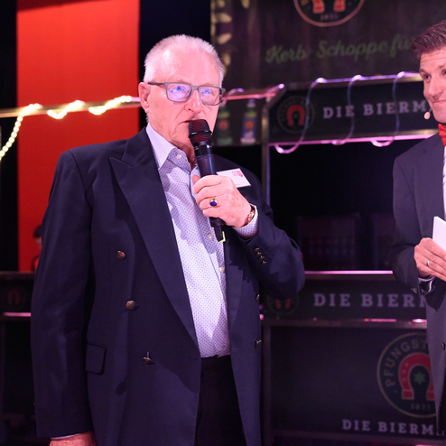 Zwei Männer stehen auf einer Bühne, einer spricht ins Mikrofon, der andere hält Notizen und hört aufmerksam zu.