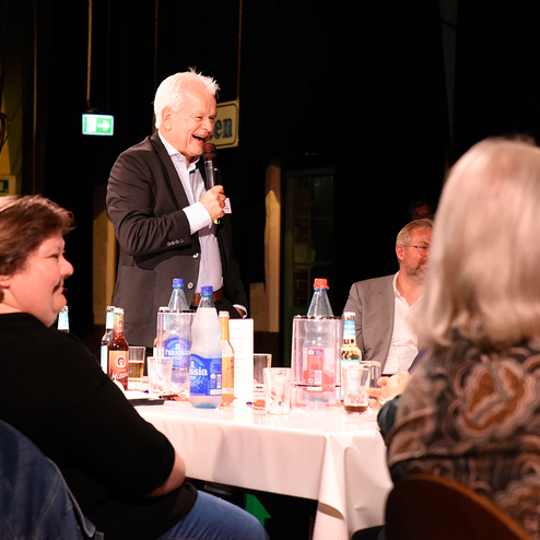 Ein Redner spricht mit Mikrofon vor einem Tisch, umgeben von Gästen in einem festlichen Ambiente.