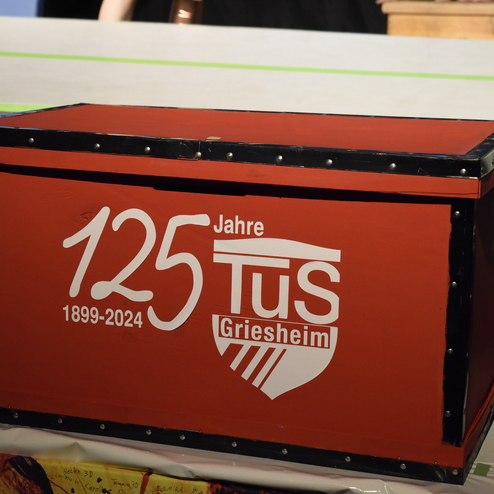 Rote Holzkiste mit der Aufschrift "125 Jahre TuS Griesheim 1899-2024" steht auf einer Tischtennisplatte.