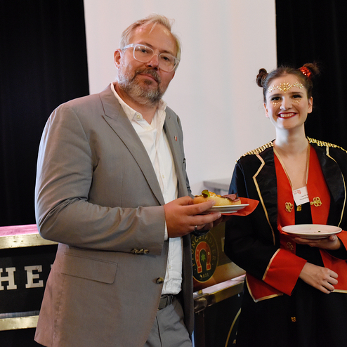 Ein Mann in einem grauen Anzug und eine Frau in einem roten Kostüm halten Teller mit Snacks bei einer Veranstaltung.