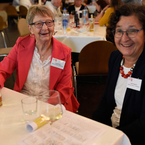 Zwei lächelnde Frauen sitzen an einem Tisch mit Getränken und Blick auf eine festliche Veranstaltung im Hintergrund.