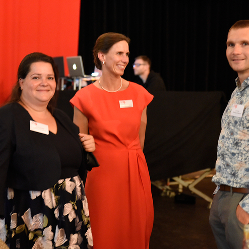 Drei Personen stehen lächelnd in einem Veranstaltungsraum, hinter ihnen rotes und schwarzes Dekor.