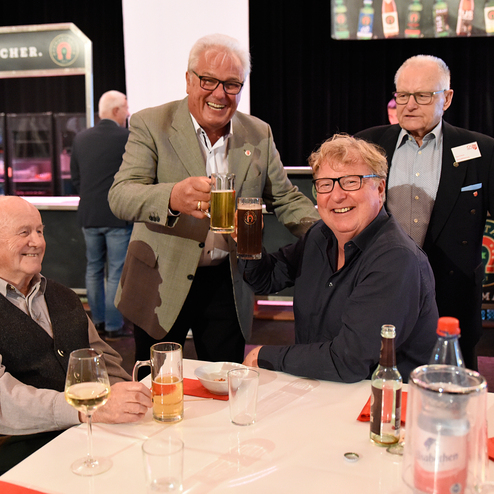 Vier Männer heben ihre Biergläser an einem Tisch, während im Hintergrund eine Getränkeausgabe zu sehen ist.