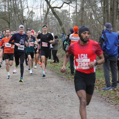 Läufer in Sportbekleidung absolvieren einen Wettkampf auf einem Waldweg, umgeben von Zuschauern und Bäumen.