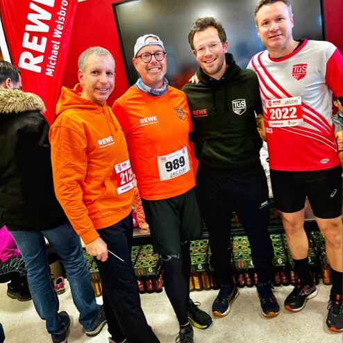 Vier Männer in Sportbekleidung mit Startnummern stehen vor einem Bildschirm in einem Laufveranstaltungsraum.