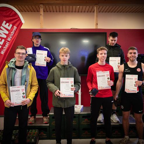 Sieben Personen halten Urkunden und stehen auf einer Plattform, im Hintergrund ein Bildschirm und REWE-Werbeflaggen.