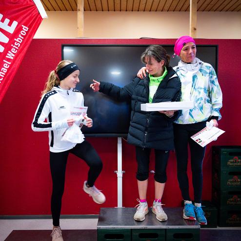 Drei Frauen stehen auf einem Podest, feiern gemeinsam und halten Urkunden und Preise in einer Sportveranstaltung.