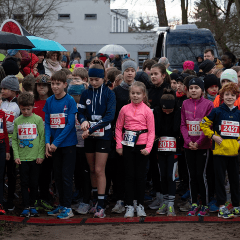Gruppenfoto von Kindern in Sportkleidung an der Startlinie eines Laufwettbewerbs, umgeben von Zuschauern.