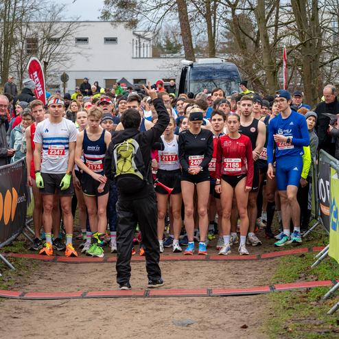Startaufstellung von Läufern bei einem Wettkampf, während ein Schiedsrichter den Beginn anzeigt. Zuschauer im Hintergrund.