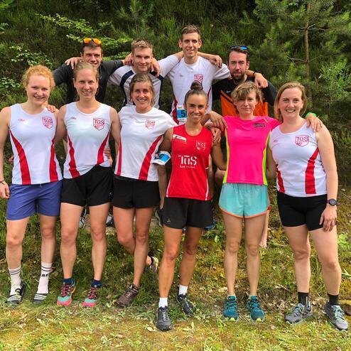 Gruppe von acht Läufern in Wettkampfkleidung steht im Freien vor einer grünen Kulisse.