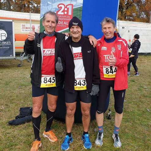 Drei Läufer geben Daumen hoch, stehen lächelnd vor einem Banner bei einem Laufereignis im Freien.