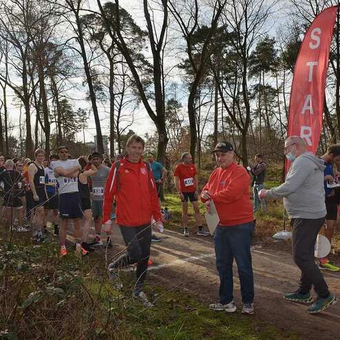 Startbereich eines Laufevents mit zahlreichen Läufern und zwei Veranstaltern im Vordergrund, umgeben von Bäumen.