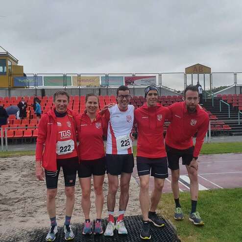 Fünf Leichtathleten in roten Trikots stehen auf einem Sportplatz vor einer Zuschauertribüne.