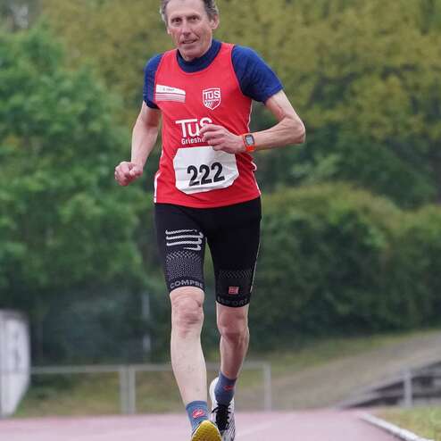 Älterer Mann in rot-blauem Trikot läuft auf einer Laufbahn, trägt die Nummer 222 und sportliche Shorts.