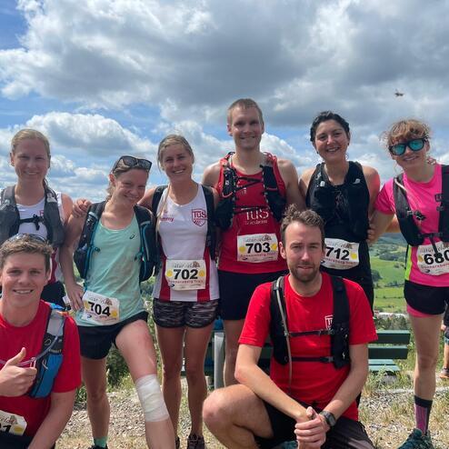 Gruppe von acht Trailrunnern mit Wettkampfnummern auf einem Gipfel, Hintergrund mit grünen Hügeln und wolkigem Himmel.