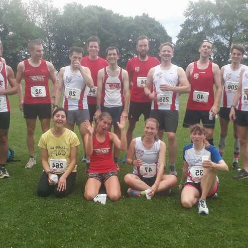 Gruppe von Läufern in Sportkleidung posiert auf einem grünen Rasenfeld nach einem Wettkampf, einige halten Becher.