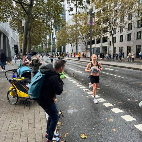 Läuferin in Sportkleidung sprintet auf regennasser Straße vorbei, während Zuschauer applaudieren und ein Kinderwagen steht.