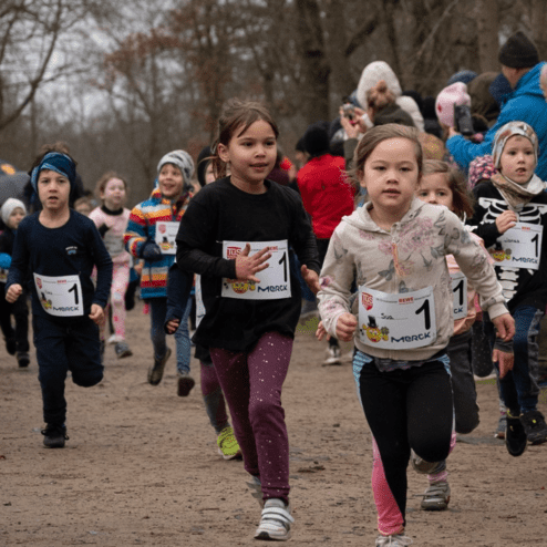 Eine Gruppe von Kindern läuft bei einem Wettkampf auf einem unbefestigten Weg, Zuschauer klatschen im Hintergrund.