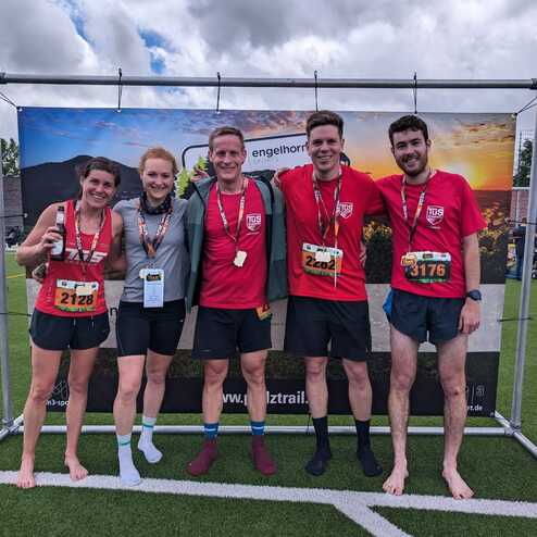 Fünf Personen in Laufbekleidung stehen vor einem Banner nach einem Wettkampf, ein paar Medaillen und Flasche in der Hand.