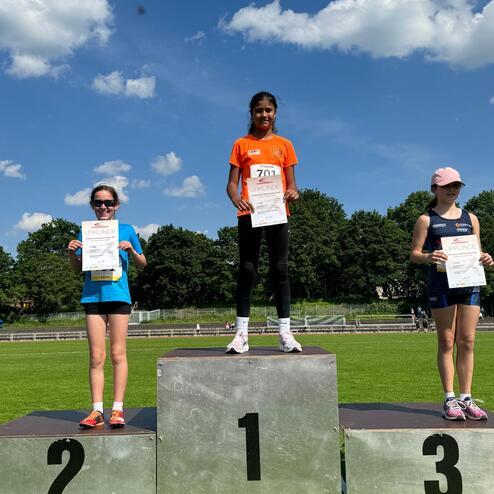 Drei Mädchen auf einem Siegertreppchen halten Urkunden, bei sonnigem Wetter unter einem blauen Himmel.