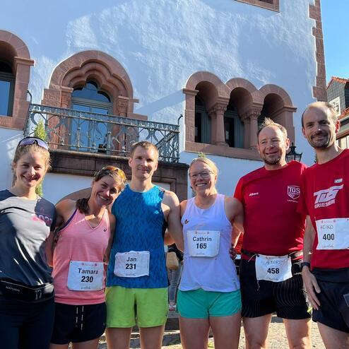 Sechs Laufsportler stehen vor einem historischen Gebäude, alle tragen Startnummern und lächeln in die Kamera.