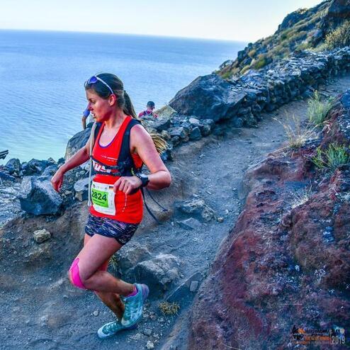 Läuferin in rotem Shirt und Shorts, die einen schmalen Pfad entlang der Küste mit Blick auf das Meer hinunterläuft.