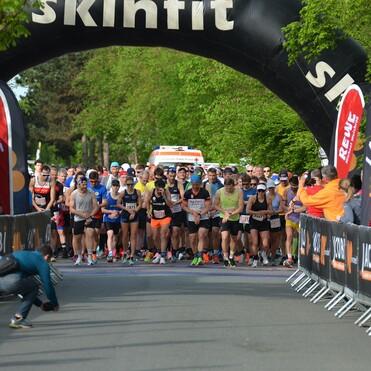 Startlinie eines Straßenrennens mit laufenden Teilnehmern und einer Uhr, die 00:00 zeigt. Banner und Zuschauer sind sichtbar.