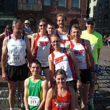 Gruppe von zehn Läufern in Sportkleidung, posierend vor einem Denkmal auf einem sonnigen Platz.