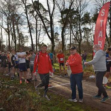 Startbereich eines Laufevents mit zahlreichen Läufern und zwei Veranstaltern im Vordergrund, umgeben von Bäumen.
