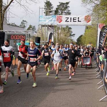 Start eines Rennens mit mehreren Läufern, einer Zeitmessung und Zuschauern entlang der Strecke. Banner mit "ZIEL".