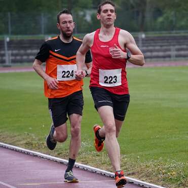 Zwei Läufer in Wettkampfbekleidung laufen auf einer Laufbahn, einer in rot mit Nummer 223, der andere in orange mit Nummer 224.