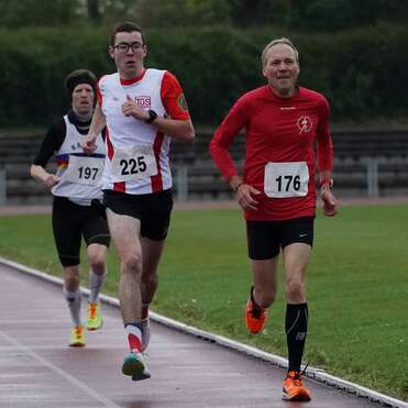 Drei Läufer in Sportkleidung sind auf einer Laufbahn unterwegs, während einer von ihnen lächelt.