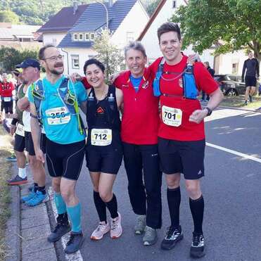 Vier Läufer stehen lächelnd zusammen, tragen Wettkampfnummern und Sportkleidung vor einem Lauf-Event in einer ruhigen Straße.