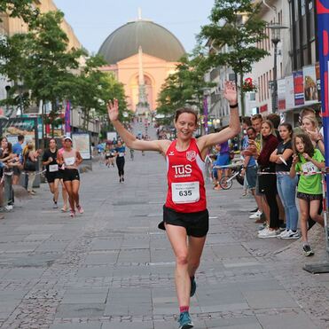Läuferin in rotem Trikot mit Händen in der Luft überquert die Ziellinie, umgeben von Zuschauern und einer Stadtansicht.