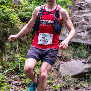 Läufer in rotem Shirt springt über felsigen Weg in einem bewaldeten Gebiet während eines Trail-Rennens.