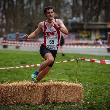 Läufer überquert einen Strohballen während eines Wettbewerbs auf einer grünen Wiese mit abgesperrtem Gelände.
