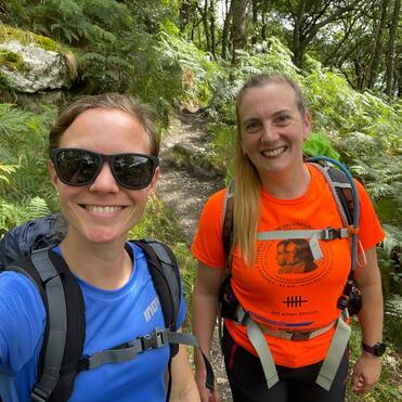 Selbstporträt zweier Frauen auf einem Wanderweg, umgeben von Farnen und Bäumen, beide tragen Wanderbekleidung und Rucksäcke.