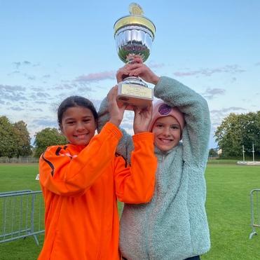 Zwei Mädchen halten triumphierend einen Pokal in die Höhe, vor einem grünen Sportfeld und einem klaren Himmel.
