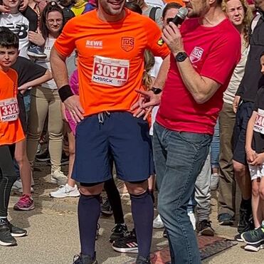 Zwei Männer sprechen auf einer Laufveranstaltung, umgeben von Zuschauern, der eine trägt ein oranges T-Shirt mit Startnummer.