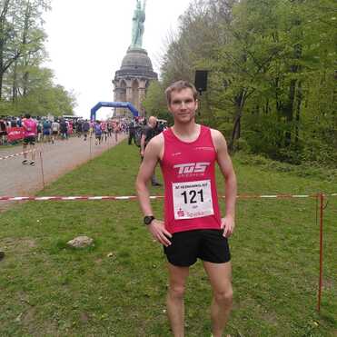 Läufer im roten Tanktop steht vor dem Hermannsdenkmal während eines Volkslaufs, umgeben von grüner Natur.