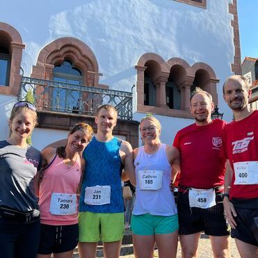 Sechs Laufsportler stehen vor einem historischen Gebäude, alle tragen Startnummern und lächeln in die Kamera.