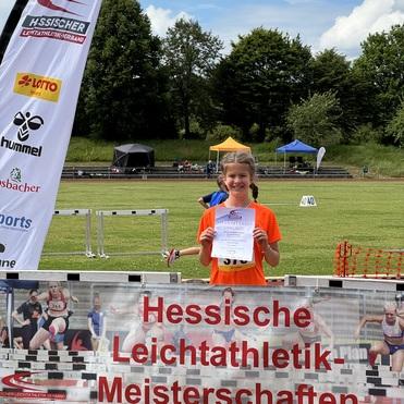Mädchen in orange T-Shirt hält Urkunde vor einem Banner der Hessischen Leichtathletik-Meisterschaften im Freien.