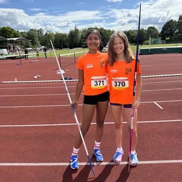 Zwei Mädchen in orangefarbenen Sportshirts stehen auf einer Laufbahn und halten Speere, im Hintergrund ein Sportplatz.