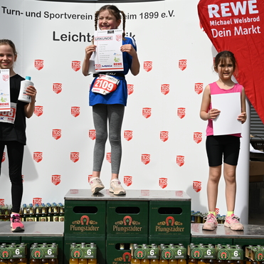 Drei Mädchen stehen auf Siegerehrungskisten, halten Urkunden und Freuen sich, im Hintergrund Banner und Getränke.