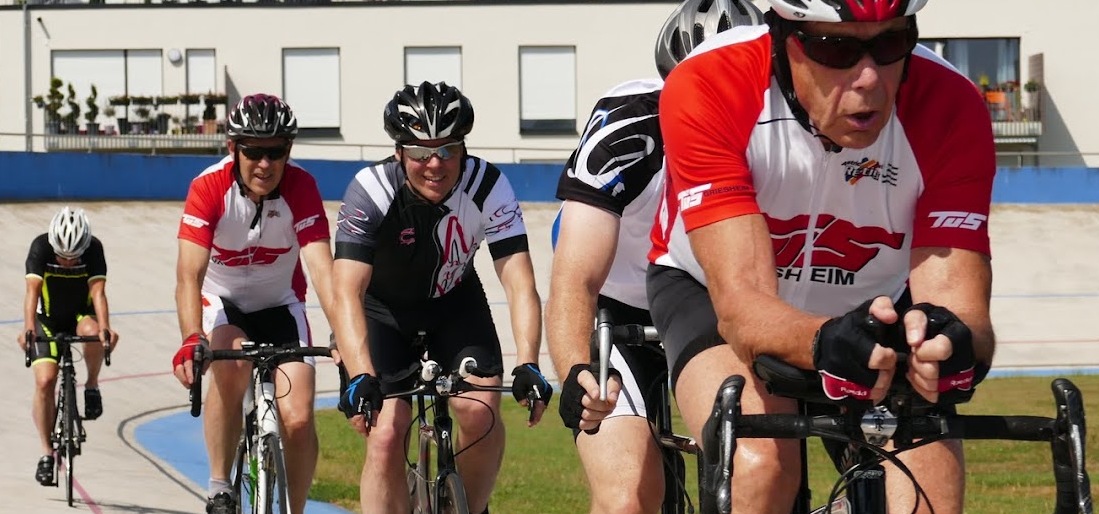 Sechs Radfahrer in Wettkampfkleidung fahren auf einer Radrennbahn mit blauer Umrandung.