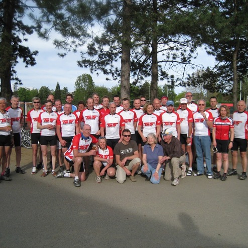 Gruppenfoto von Radfahrern in roten und weißen Trikots, stehend vor einer Sportanlage unter Bäumen.