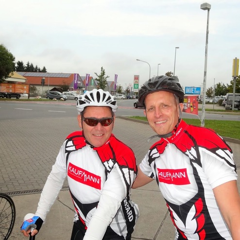 Zwei Radfahrer in rot-weißer Sportkleidung posieren lächelnd an einer Straßenecke, Fahrräder im Hintergrund.