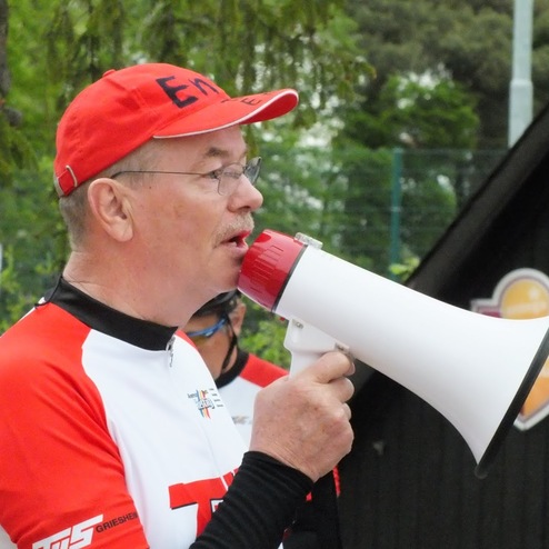Mann mit Brille und roten Kappen spricht in ein Megafon, im Hintergrund Bäume und eine dunkle Holzstruktur sichtbar.