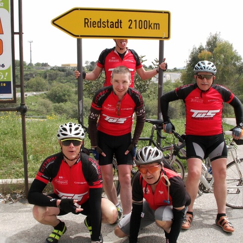 Gruppe von fünf Radfahrern in roten Trikots posiert vor einem „Riedstadt 2100km“-Schild an einer Landstraße.