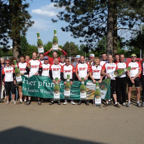 Gruppenshot von Radfahrern in festlichen Trikots, die ein Banner und große Bierflaschen gemeinsam halten.